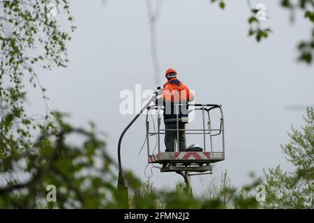An electrician in special clothing and a helmet repairs a street lamp and screws a street light bulb on a Stock Photo