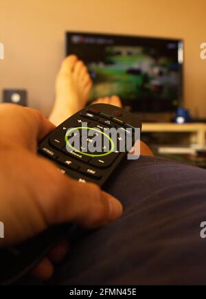 Hand of a man holding a remote control while watching tv or streaming movie or series with smart tv at home with a view of the feet. Stock Photo