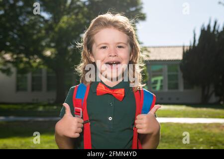 Cheerful little school boy in school uniform with big backpack standing near school. Back to School. Kids preschool education concept. Stock Photo