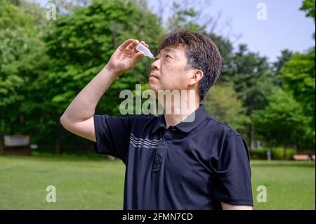Middle-aged Asian man putting eye drops in his eyes outdoors. Stock Photo