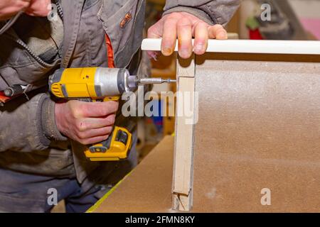 Worker in carpentry uses professional manual power tool, acu drill, and twists the screws on the wooden door frame. Stock Photo