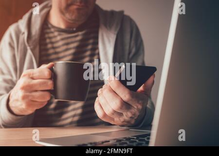Freelance worker using mobile smart phone and drinking coffee at home office desk, close up with selective focus Stock Photo