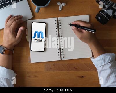 Overhead shot of male office worker using mock up smartphone while taking note on blank notebook on wooden table Stock Photo