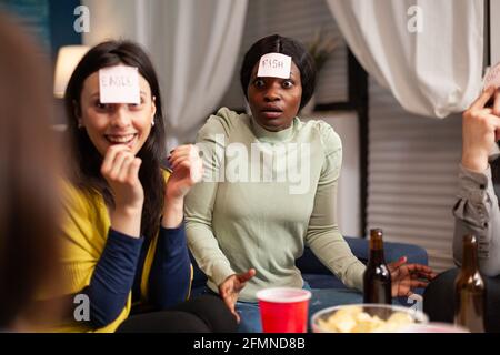 Group of multi-ethnic people having fun while playing board game using sticky notes spending time together. Creative friends hanging out late at night in living room during home party. Stock Photo
