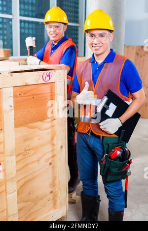 Two Asian Indonesian industrial or construction workers controlling with a checklist a delivery on a tower building site and open a wood box or cargo Stock Photo