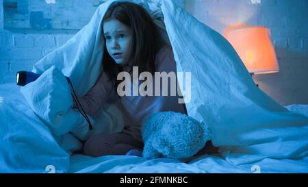 Scared child holding flashlight near teddy bear under blanket on bed Stock Photo