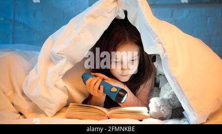 Preteen kid holding flashlight while reading book near soft toy on bed Stock Photo