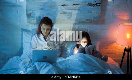 Freelancer using laptop near daughter with digital tablet in bedroom in night Stock Photo