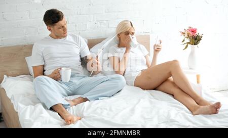 tattooed man reading newspaper near woman drinking coffee and using smartphone in bed Stock Photo