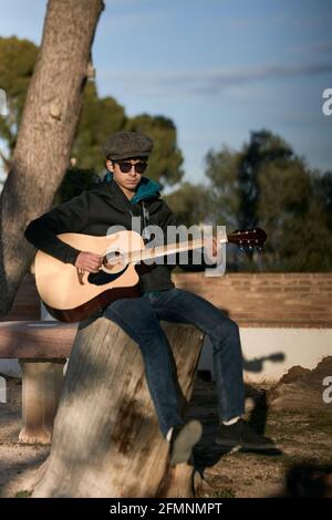young man sitting in attractive attire playing the acoustic guitar outdoors Stock Photo