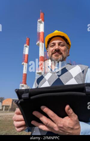 Smiling Businessperson in Yellow Hardhat Holding Digital Tablet and Standing Against Power Plant Chimneys and Blue Sky Background. Stock Photo