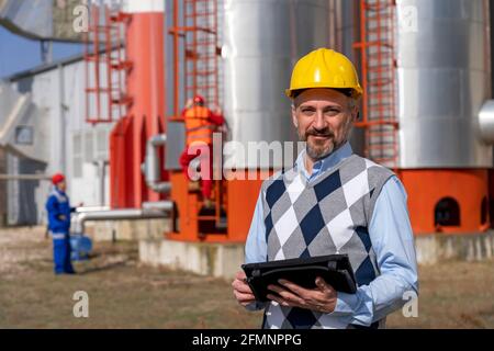 Oil Engineer With Digital Tablet Standing in front of Oil Refinery Storage Tanks. Portrait of Businessperson in Yellow Work Helmet. Stock Photo