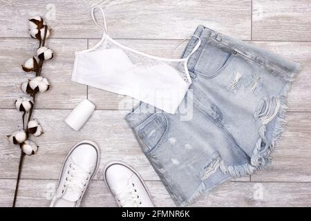Summer fashion women's outfit. Blue ripped denim shorts, white bra, sneakers, roll-on deodorant with cotton sprig. Summer essentials Stock Photo
