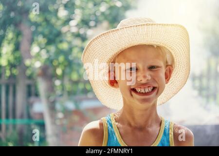 Cheerful little boy wearing large straw hat and blue sleeveless shirt posing for camera and smiles all over his mouth in sunny summer city park close Stock Photo