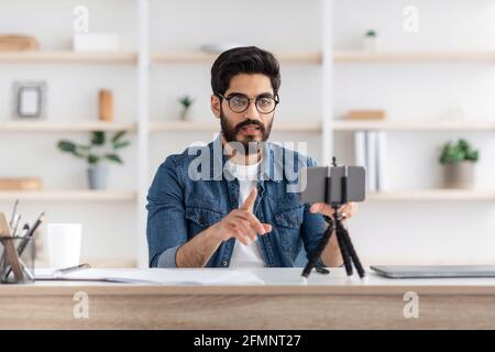 Arab man broadcasting from home, using smartphone and tripod, sitting at table and talking to phone camera Stock Photo