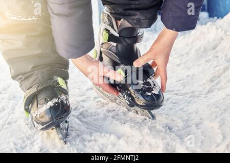 Boy in warm jacket and pants adjusts ice skates standing on snowy ground near rink on cold day extreme close view Stock Photo