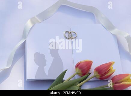 Summer wedding mockup. Blank wedding book with a silhouette of lovers on a white table. Wedding rings are placed on the book. Stock Photo