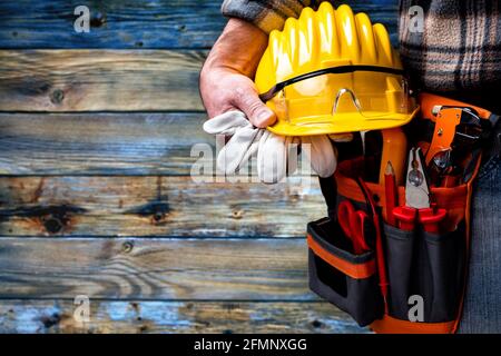 Electrician worker on vintage light blue wooden background; holds helmet, gloves and goggles in hand. Construction industry, electrical system. Stock Photo