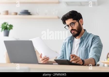 Expensive modern life. Disappointed arab man calculating taxes or planning budget while sitting at kitchen table Stock Photo