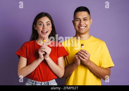 Brunette happy couple smiling while posing with lollipops isolated over purple wall Stock Photo
