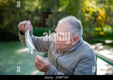 old gray-haired man puts on a medical mask. Prevention of covid 19 infection for the elderly. A man on the street takes off his medical mask due to th Stock Photo