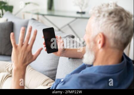 Grey haired senior middle-aged man using smartphone for video call, phone with black empty screen in his hand, back view, mature man waving hello to blank screen, video call, virtual meeting. Mockup Stock Photo