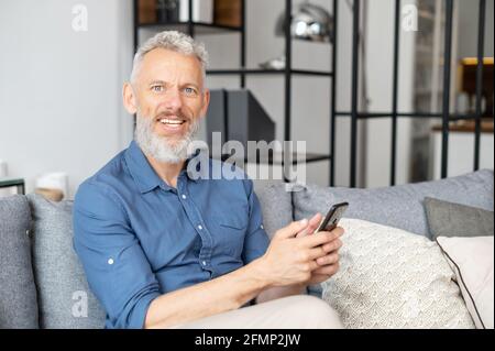 Cheerful and healthy grey-haired middle aged hipster man in casual shirt using smartphone sitting on the sofa at home, mature guy enjoys online chatting, looks at the camera and smiles Stock Photo