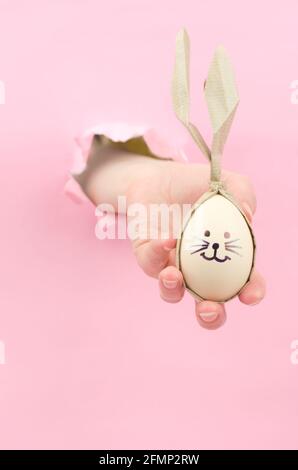 Easter eggs in the form of a rabbit in a female hand on a pink background. Eggs with painted bunny faces, easter concept. Preparations for the Easter Stock Photo