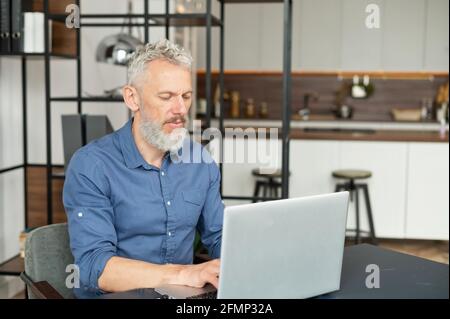 Focused mature man typing on the keyboard, senior male using laptop for remote work, messaging, working on the new startup sitting at the desk in modern apartment Stock Photo