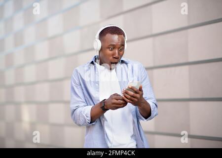 Shocked African American man in headset staring at smartphone screen on city street Stock Photo