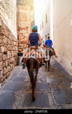 Tourists riding donkeys equipped with leather saddles on weathered narrow paved road on Lindos historical town street in Greece Stock Photo