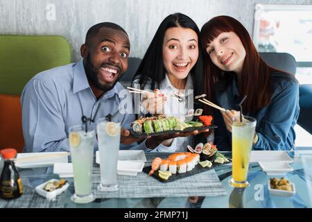 Three joyful multiracial friends, eating sushi rolls, using bamboo sticks. Young diverse people enjoying tasty asian meal in fashionable restaurant Stock Photo