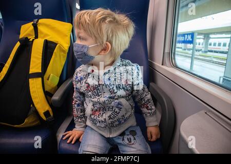 Blond kid with blue eyes and face mask, near a yellow backpack, on board a regional train in Italy Stock Photo