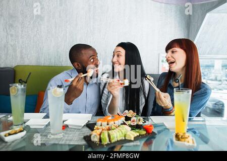 Happy three multiracial friends enjoying sushi at a Japanese restaurant. Pretty women and handsome man, eating sushi rolls and communicating, sitting at cafe Stock Photo