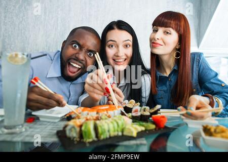 People, food and friendship concept. Three happy young friends enjoying sushi at a Japanese restaurant. Happy students eating sushi rolls and laughing at camera Stock Photo
