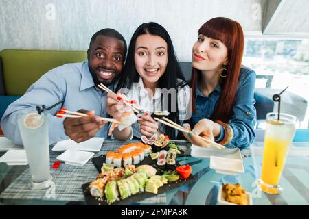 Happy three multiracial young people, posing at camera in asian restaurant, enjoying their lunch time with tasty delicious sushi rolls. Japanese food and friendship concept Stock Photo