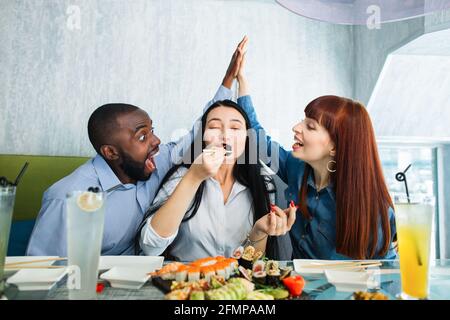 Tasty food, the pleasure of eating. Pretty young Asian woman enjoying tasty sushi roll, sitting at the table at cafe, while her friends, African man and Caucasian woman giving high five each other Stock Photo