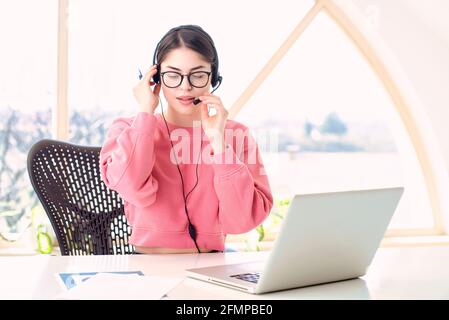 Young businesswoman in headset speaking by conference call while sitting behind her laptop. Home office. Stock Photo