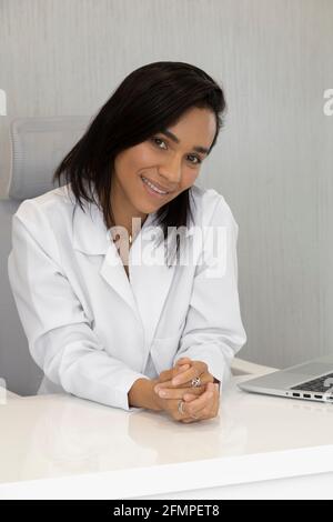 doctor's office, there is a beautiful young latin healthcare professional with short hair sitting on the desk, medicine Stock Photo