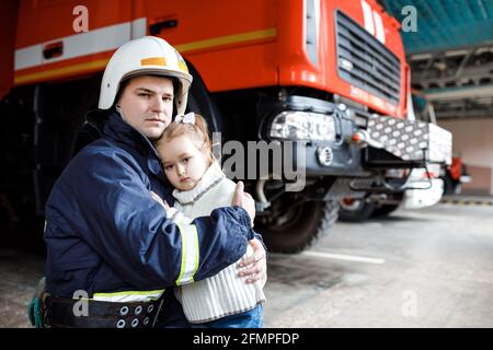 Brave firefighter in uniform holding little saved girl standing on black background Stock Photo