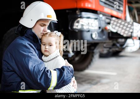 Brave firefighter in uniform holding little saved girl standing on black background Stock Photo