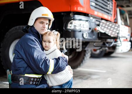 Brave firefighter in uniform holding little saved girl standing on black background Stock Photo