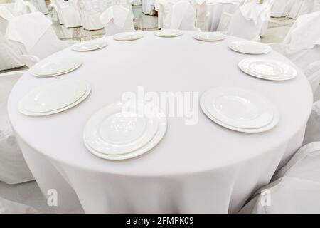 Empty white plates on a white tablecloth on a restaurant circle table. Restaurant table setting. Stock Photo