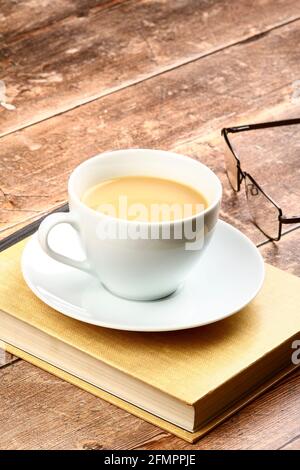 White cup and saucer filled with tea sat on a reading book on a rustic wooden table top Stock Photo