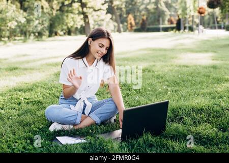 student girl with laptop outdoors. Smiling woman sitting on the grass with a computer, surfing the Internet or preparing for exams. Technology, educat Stock Photo