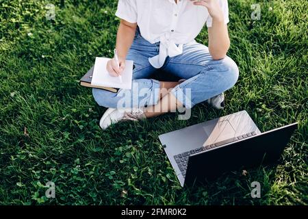 student girl with laptop outdoors sitting on the grass, surfing the internet or preparing for exams. Technology, education and remote work concept. So Stock Photo