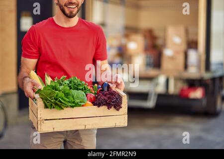 Male courier in uniform holding grocery box with fresh vegetables for delivering while standing against warehouse building Stock Photo