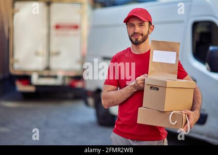Young handsome caucasian male courier in red uniform holding parcels and paper bag for delivery and looking at camera while standing at storehouse Stock Photo