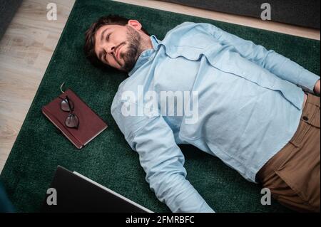 Young cute office worker lies on the floor mat next to the sofa, he is tired and asleep, top view. Stock Photo