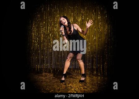 Full size photo of brown optimistic lady stand sing in mic wear black dress shoes isolated on dark gold color background in stage light Stock Photo